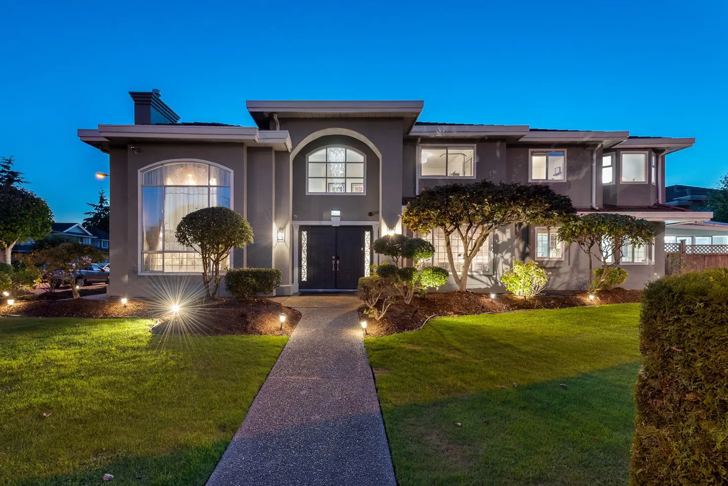 A two-story grey house at twilight with a lit walkway, manicured lawn, and exterior landscape lighting.