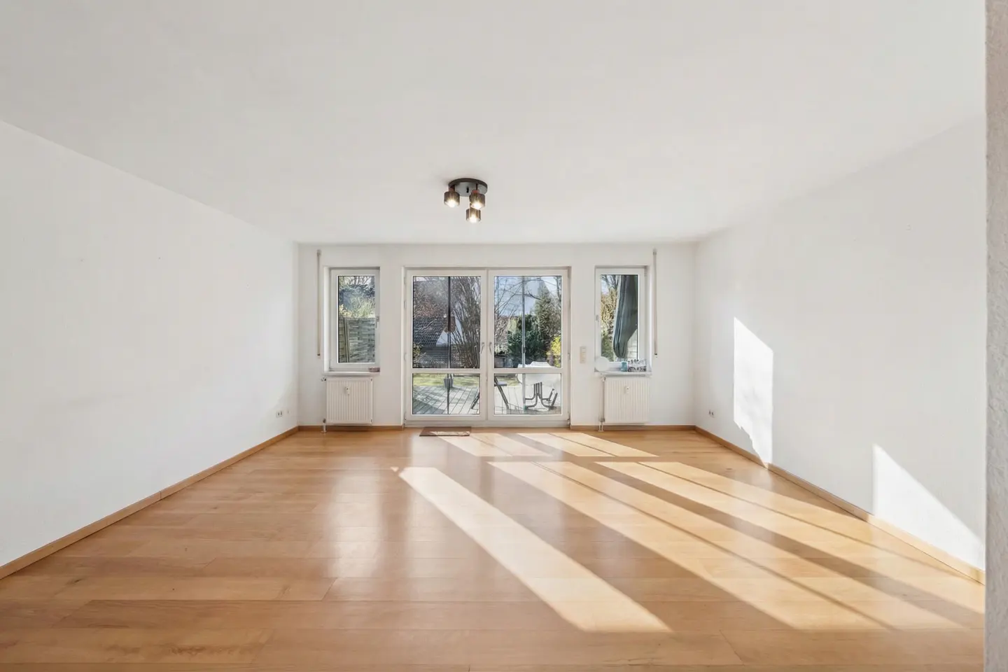 Bright, empty room with light wood floors, white walls, and a sliding glass door to a patio. Sunlight streams in.