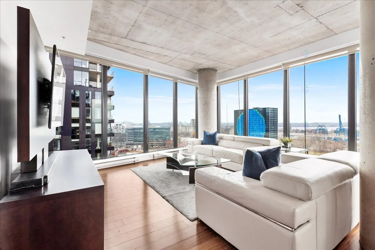 Bright living room with floor-to-ceiling windows, white sofas with blue pillows, and a glass coffee table. City view in the background.