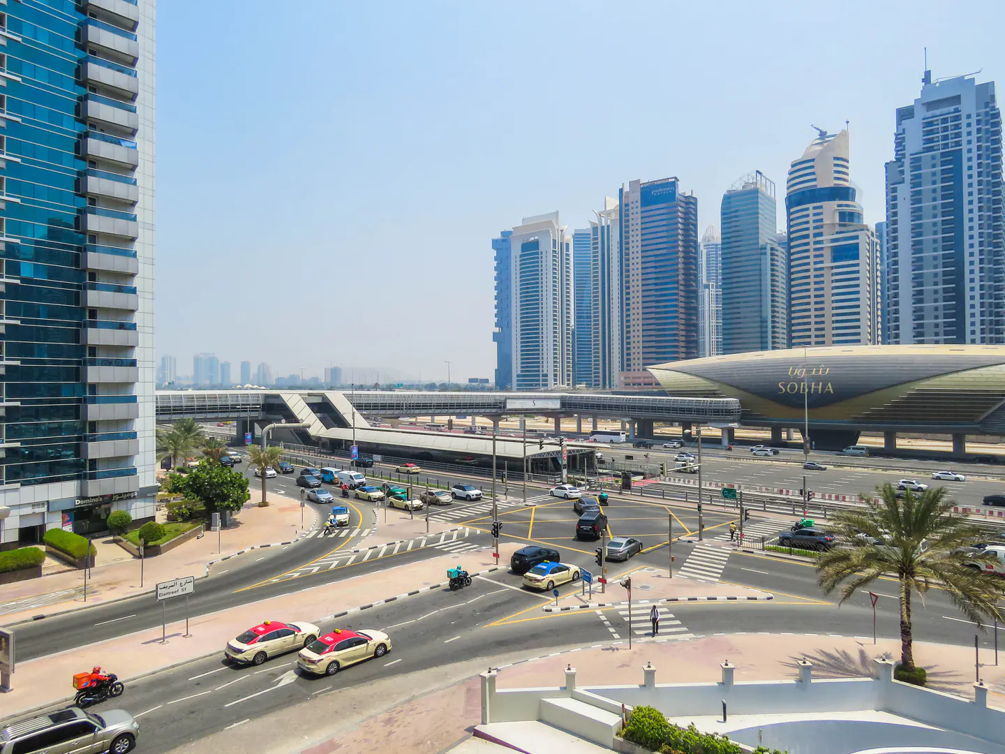 Cityscape view of Dubai with modern buildings, a metro station, and busy streets filled with cars and taxis.