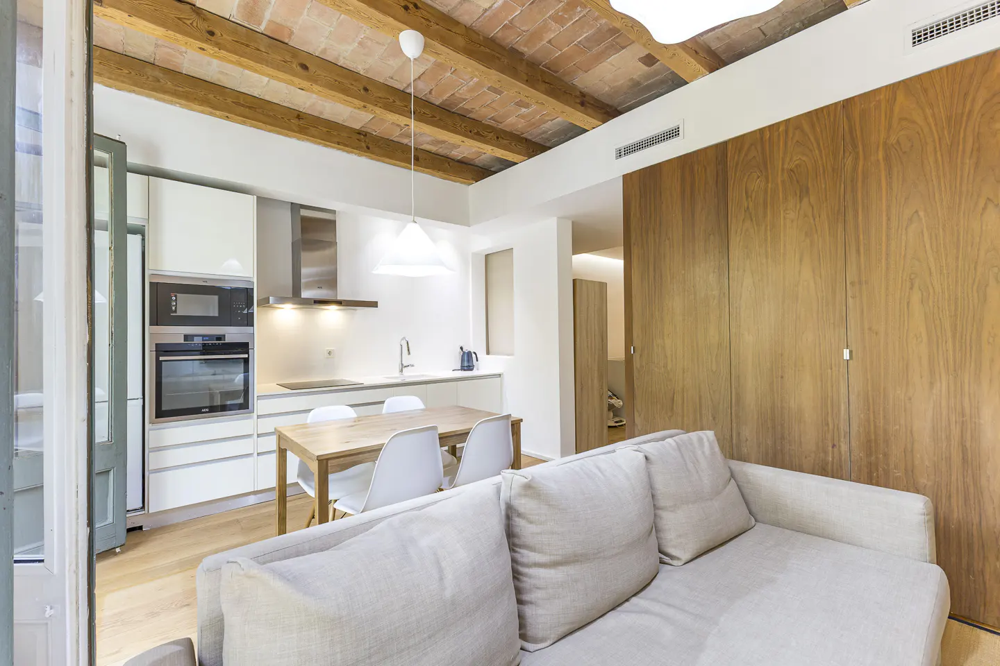 Bright apartment interior with exposed brick ceiling, white kitchen, wood table with white chairs, and a light gray sofa.