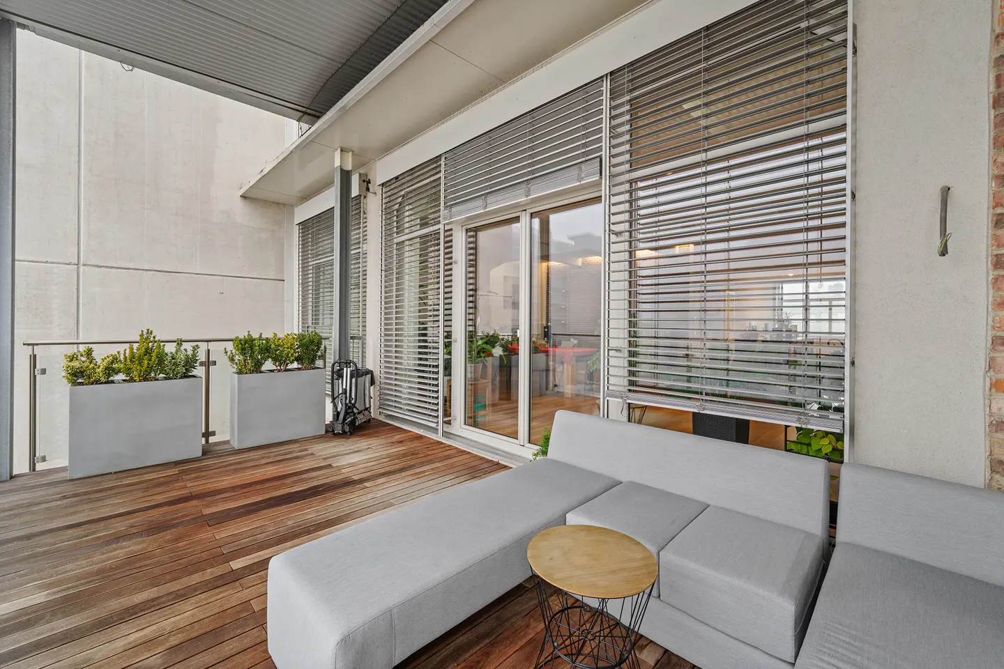 A modern balcony with a wooden floor, gray modular sofa, and round table. Planters with greenery line the railing, and blinds cover the sliding glass doors.