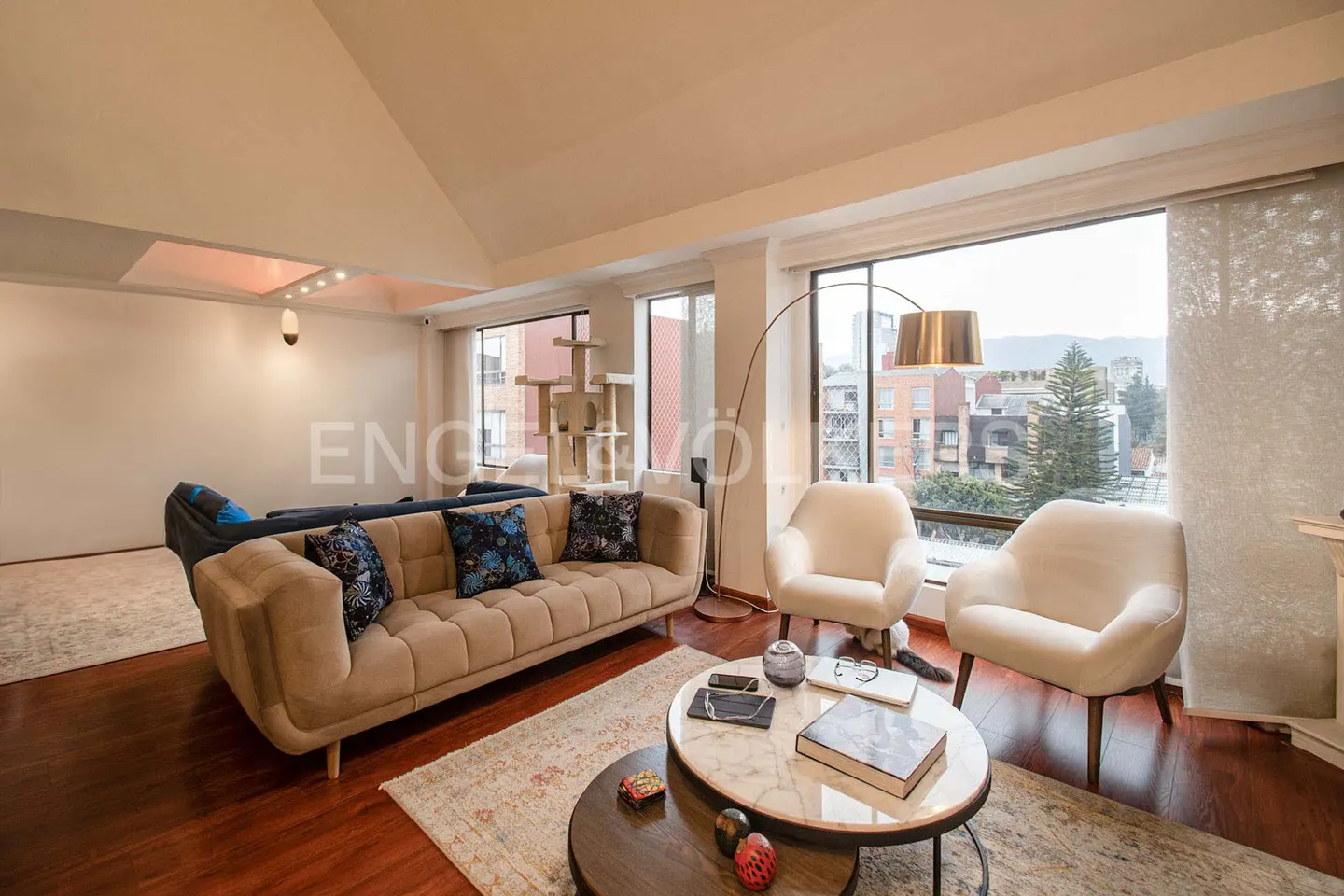 Living room with beige sofa, two white chairs, and round coffee tables. Large windows offer a city view.