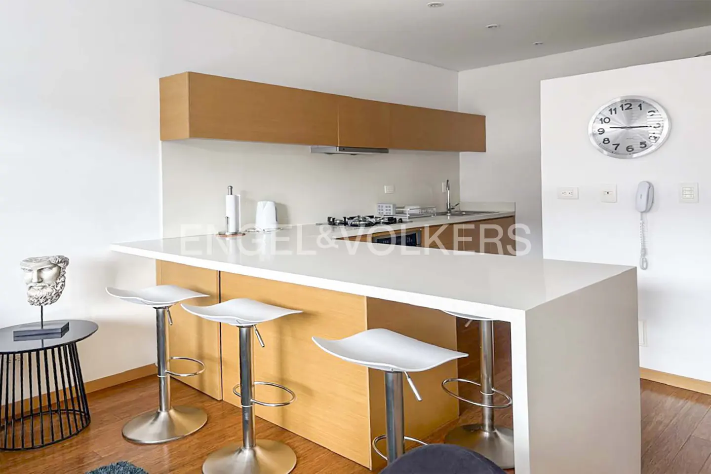 A modern kitchen with a white countertop island and three white bar stools. A clock hangs on the white wall.