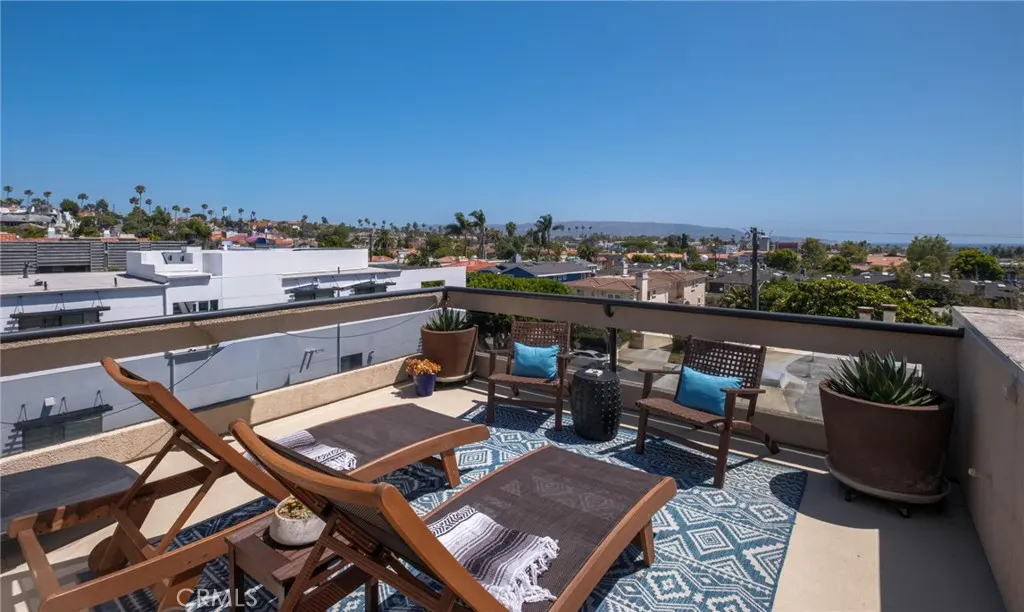 Rooftop deck with wooden lounge chairs, blue patterned rug, potted plants, and a panoramic view of a suburban neighborhood.