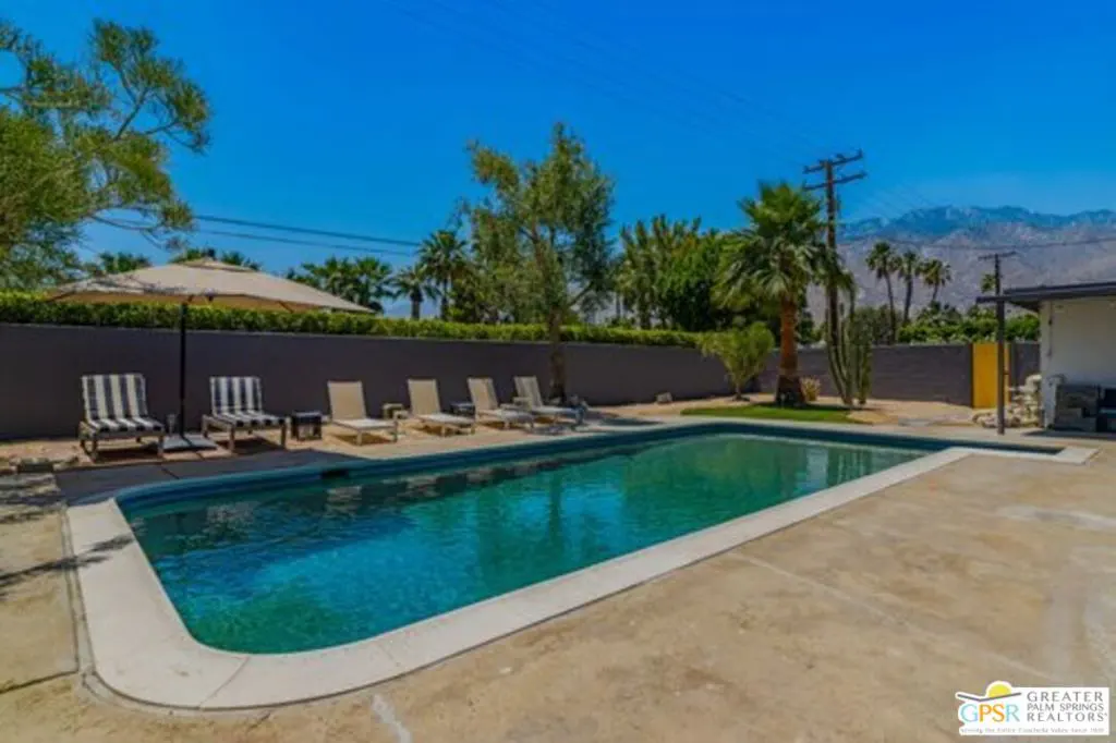 A backyard pool with lounge chairs and a tan umbrella under a clear blue sky.