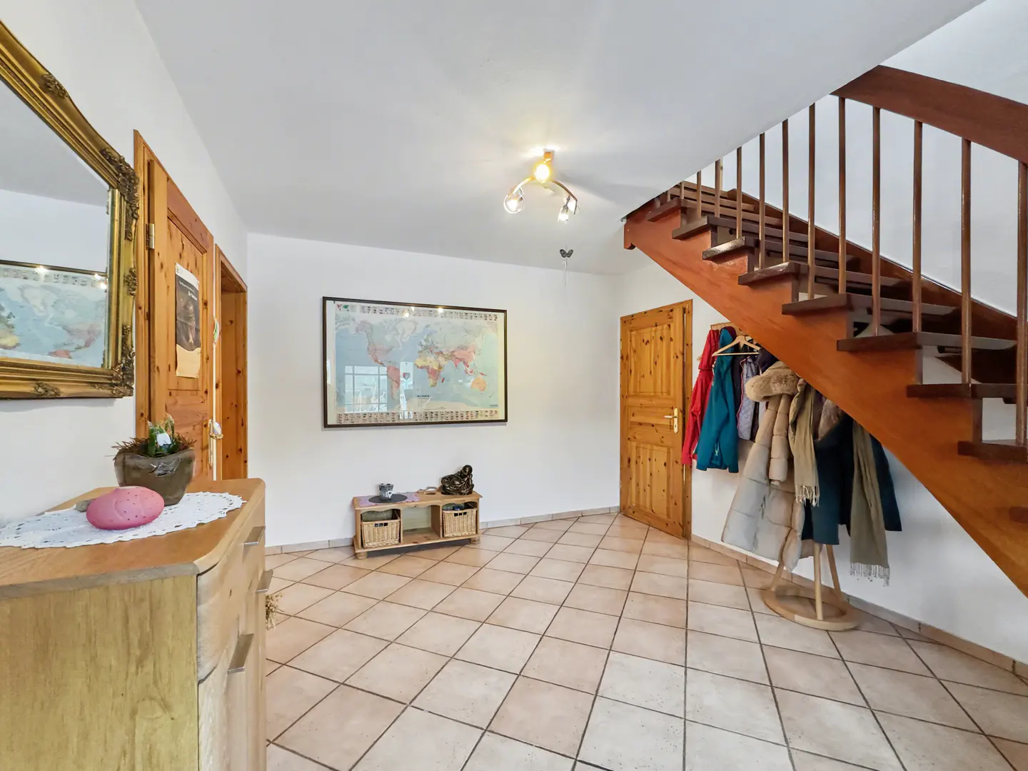 A bright foyer with tile floors, a wooden staircase, and a world map on the wall. A coat rack holds jackets near a wooden door.