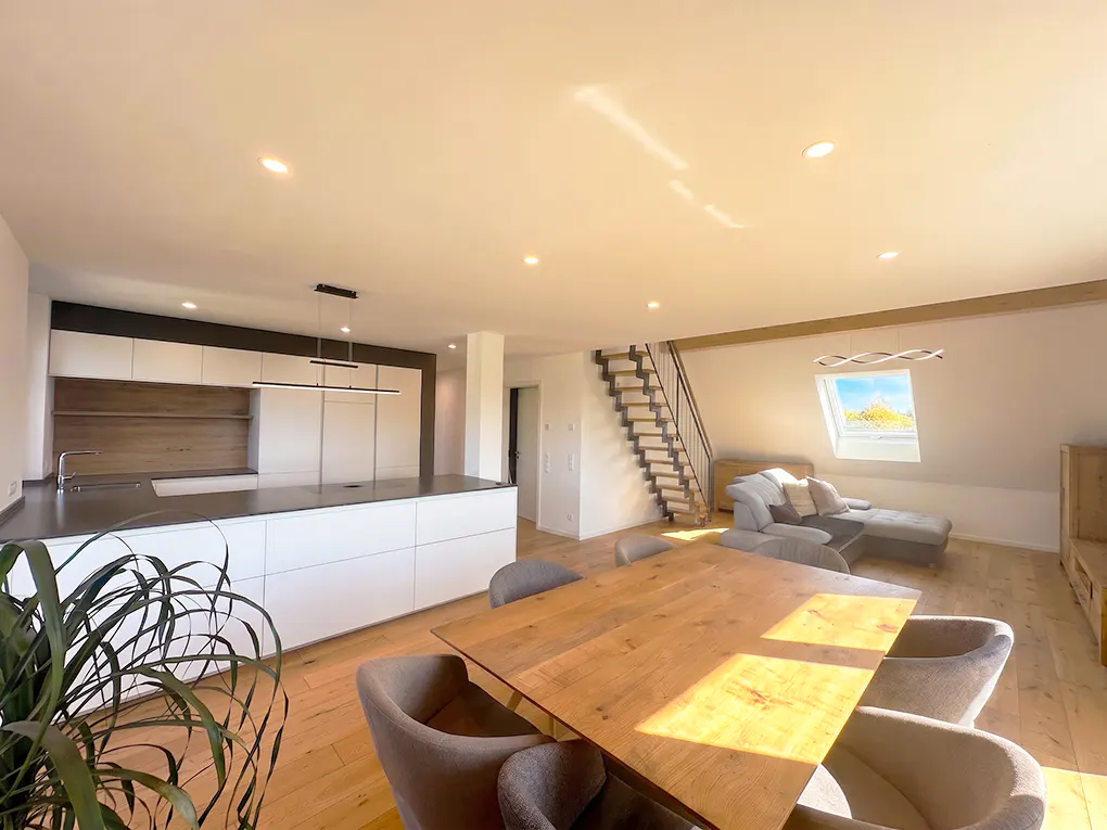 Modern open-plan living area with a white kitchen, wooden dining table, gray chairs, and a staircase leading to a loft.