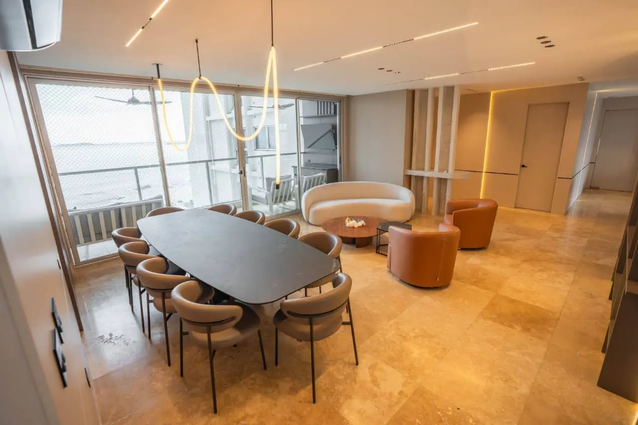 Bright, modern living space with a large dining table, curved white sofa, and tan leather chairs. Ocean view visible through large windows.