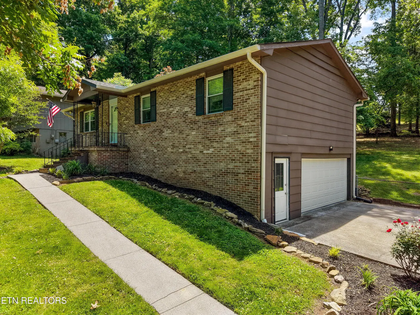 A tan brick and brown siding house with green shutters, a white garage door, and a sidewalk leading to the front door.