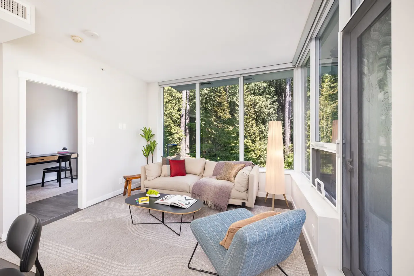 Bright living room with beige sofa, blue chair, and large windows overlooking a forest. A black coffee table sits on a patterned rug.