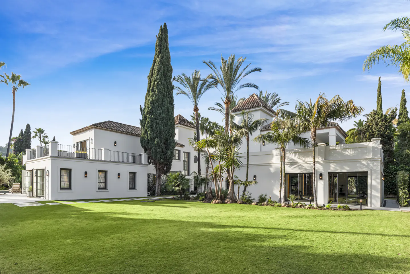 Exterior view of a large white house with a green lawn, palm trees, and a blue sky.