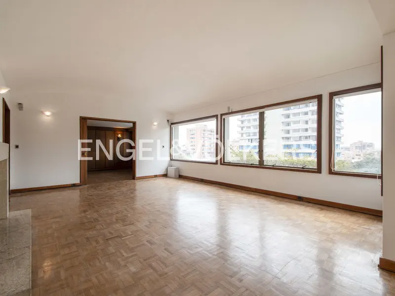 Bright, empty living room with parquet floors, white walls, and large windows with city views. A doorway leads to another room.