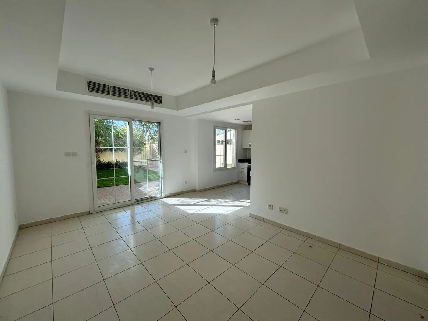 Bright, empty room with white walls and tile floor. Sliding glass doors lead to a green lawn. Kitchen visible in the background.