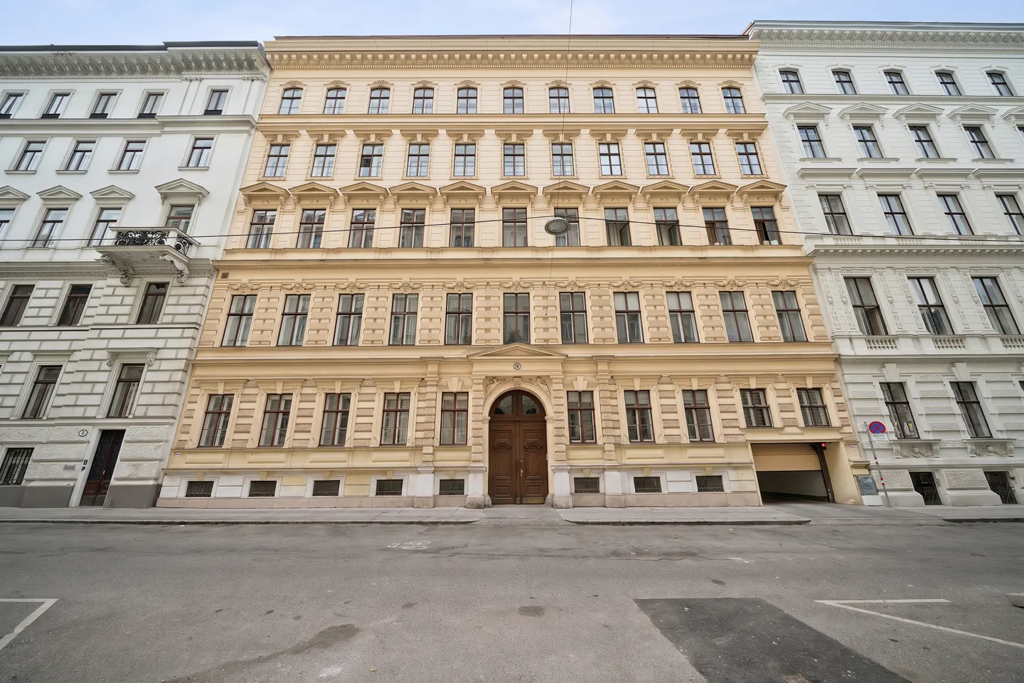 A tan, multi-story building with many windows and a large wooden door, flanked by white buildings, on a gray street.