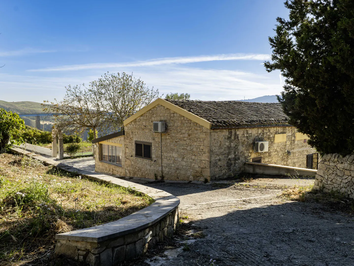 Exterior view of a stone house with a dark roof, surrounded by trees and greenery under a blue sky. A bridge is visible in the background.