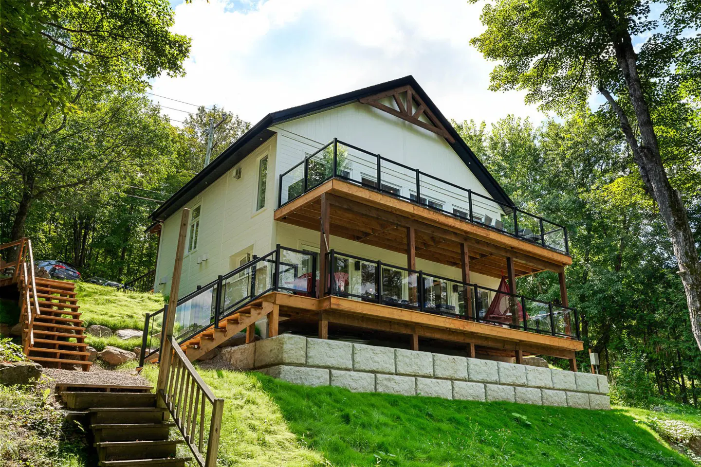 Exterior view of a two-story white house with a black roof and two wooden decks with glass railings, surrounded by green trees and grass.