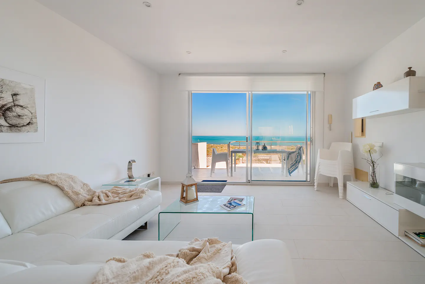 Bright, all-white living room with a glass table, white sofa, and a balcony overlooking the ocean.