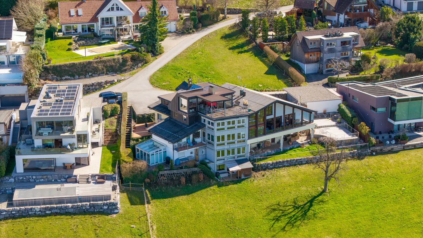 Aerial view of a multi-story house with many windows on a green lawn, surrounded by other houses and trees.