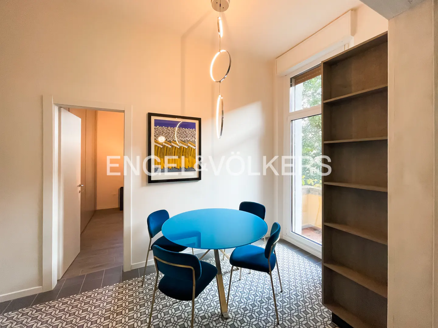 A dining area with a blue table, velvet chairs, and a modern chandelier. A bookshelf stands near a window, and a doorway leads to another room.
