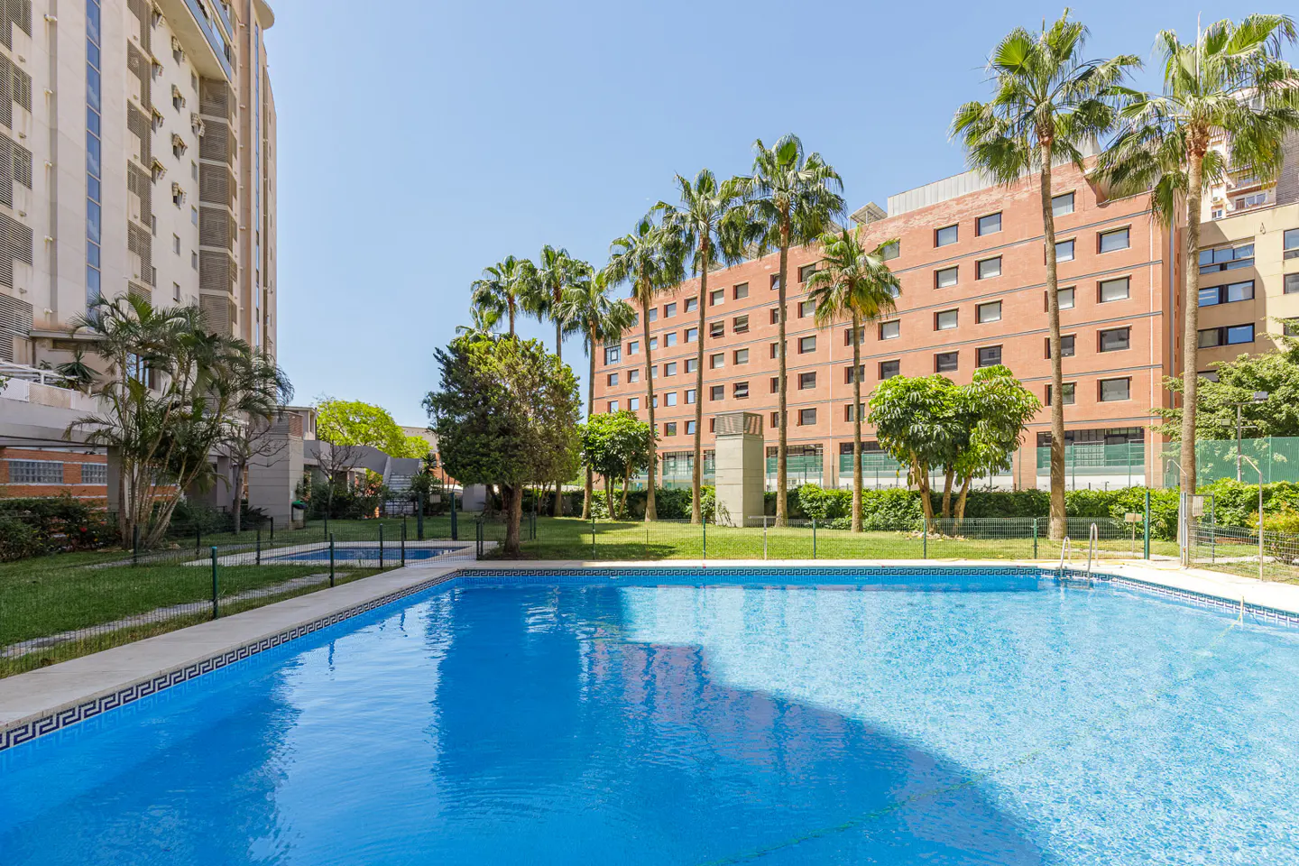 A bright blue swimming pool is surrounded by palm trees and apartment buildings on a sunny day.