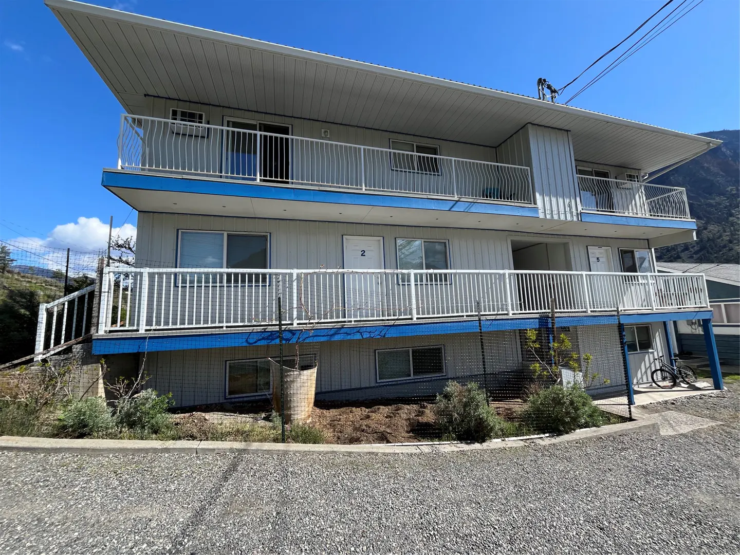 Two-story apartment building with white siding, blue trim, and white railings. Balconies on both floors. Numbered doors. Blue sky.