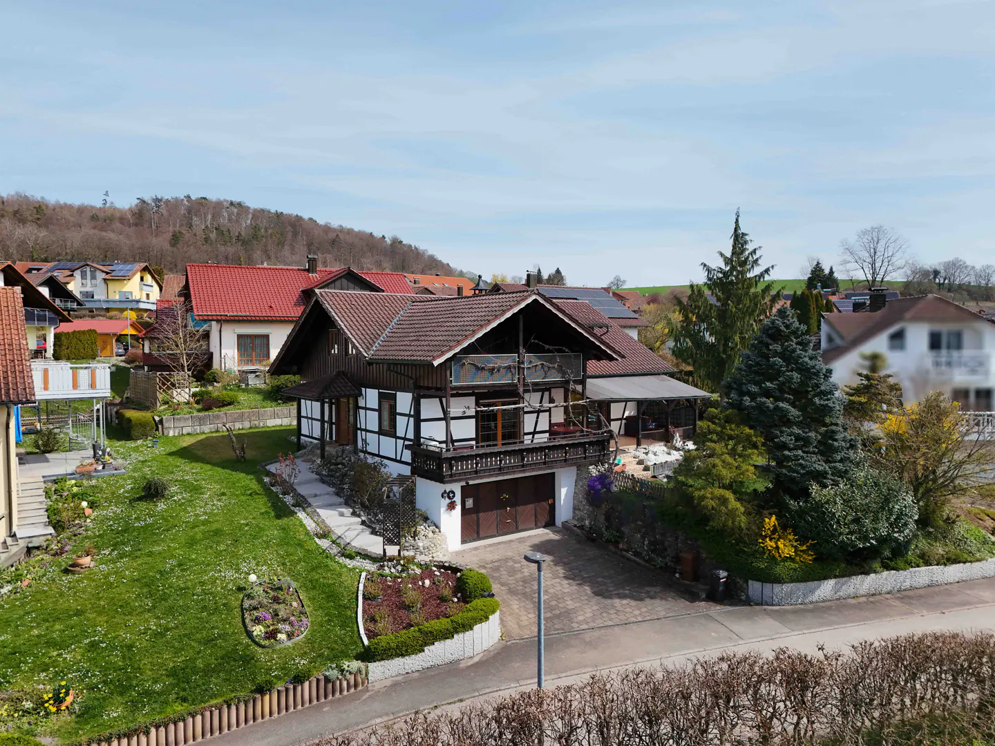 Two-story, half-timbered house with a brown roof, white walls, and a balcony, surrounded by a green lawn and trees.