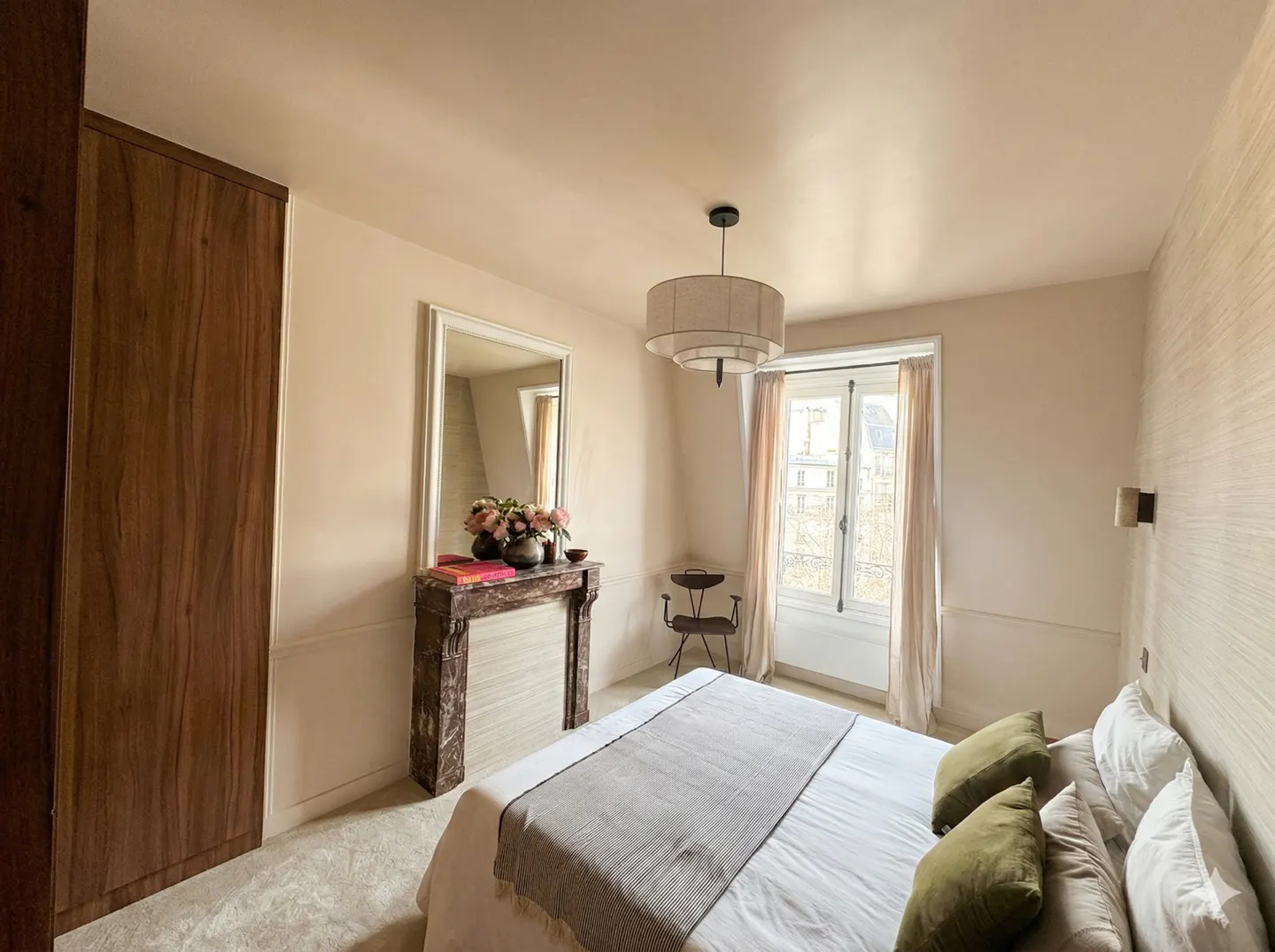 Bedroom with a bed, green pillows, a marble fireplace with a mirror, a wooden wardrobe, and a window with light curtains.