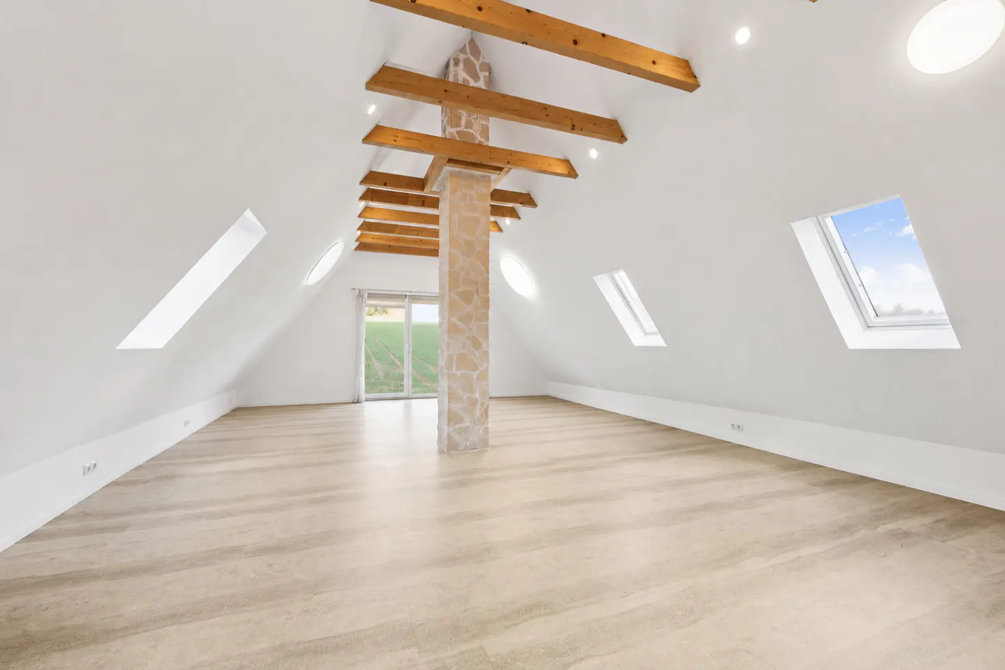 Attic room with white walls, light wood floors, and exposed beams. Skylights and a stone-clad pillar add character. Sliding glass doors lead to a green lawn.
