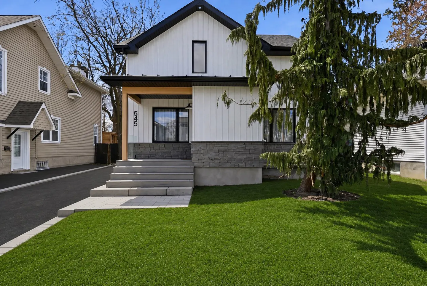 Exterior of a two-story house with white siding, black trim, and a gray stone foundation. A green lawn and a large tree are in the foreground.