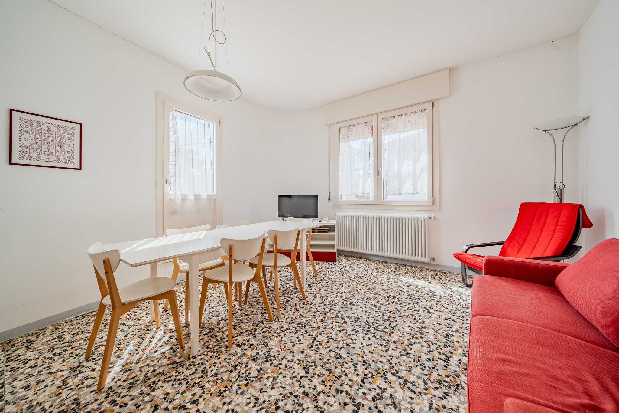 Bright living room with white walls, terrazzo floor, red sofa, white table with chairs, and two windows with white curtains.