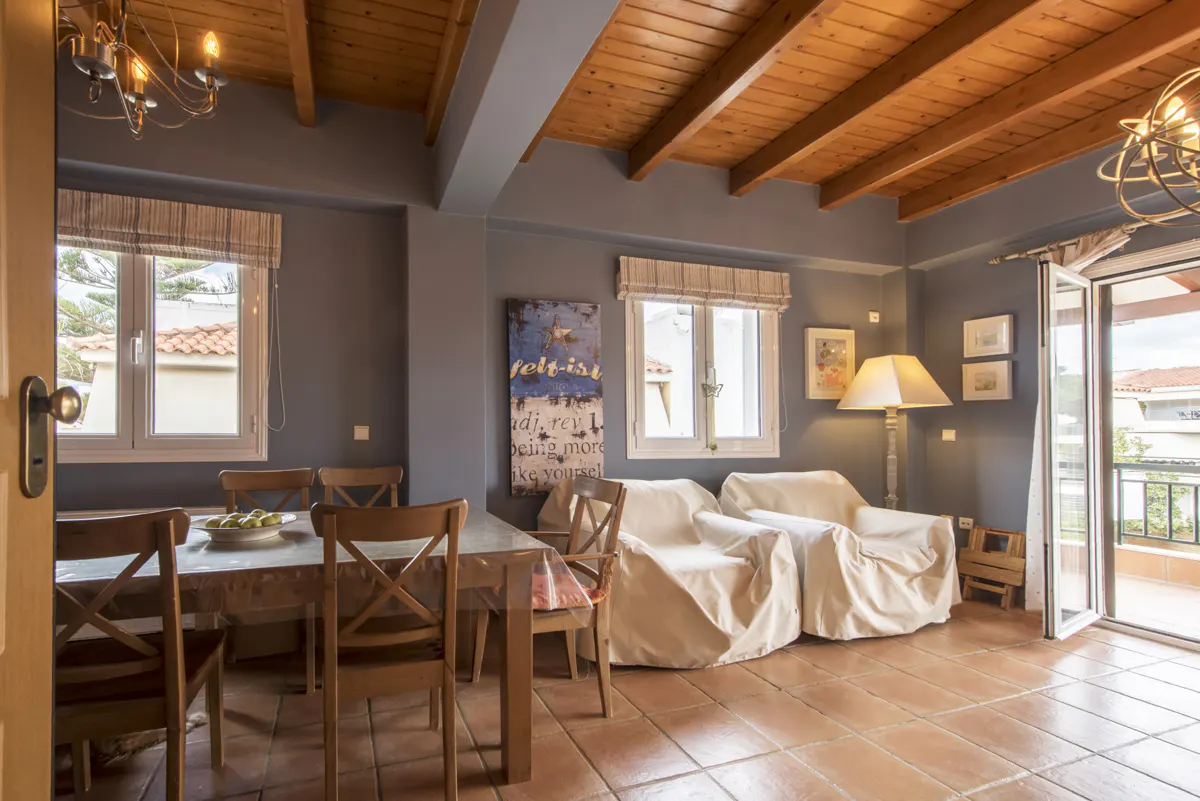 A living room with a wooden ceiling, gray walls, and terracotta tile floors. A table with chairs and covered armchairs are visible.
