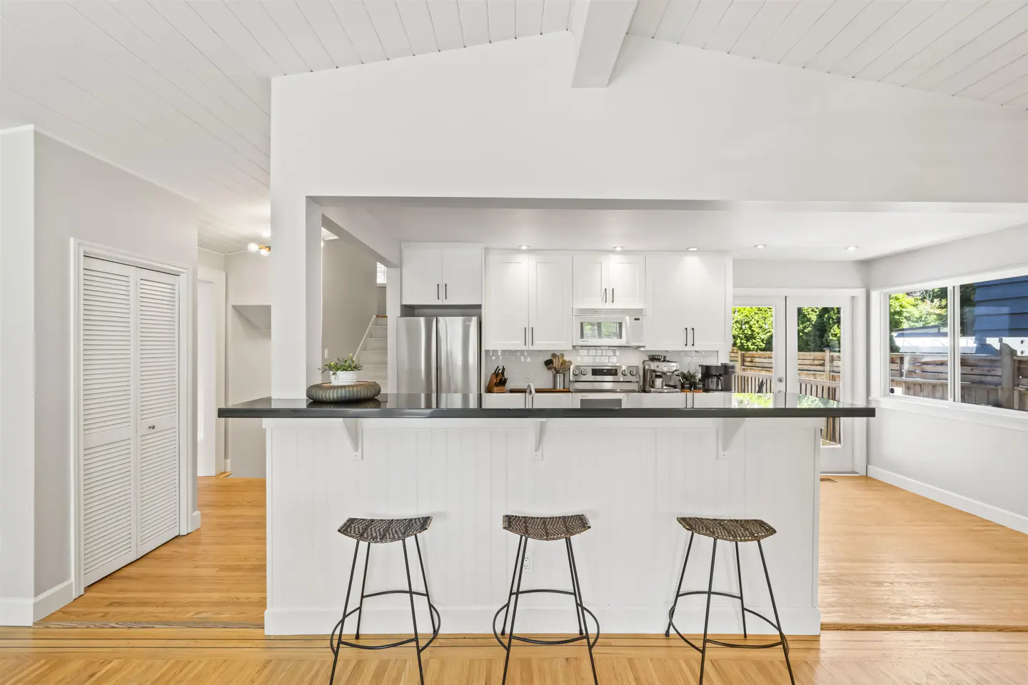 Modern kitchen with white cabinets, stainless steel appliances, black countertop, and three bar stools on hardwood floors.