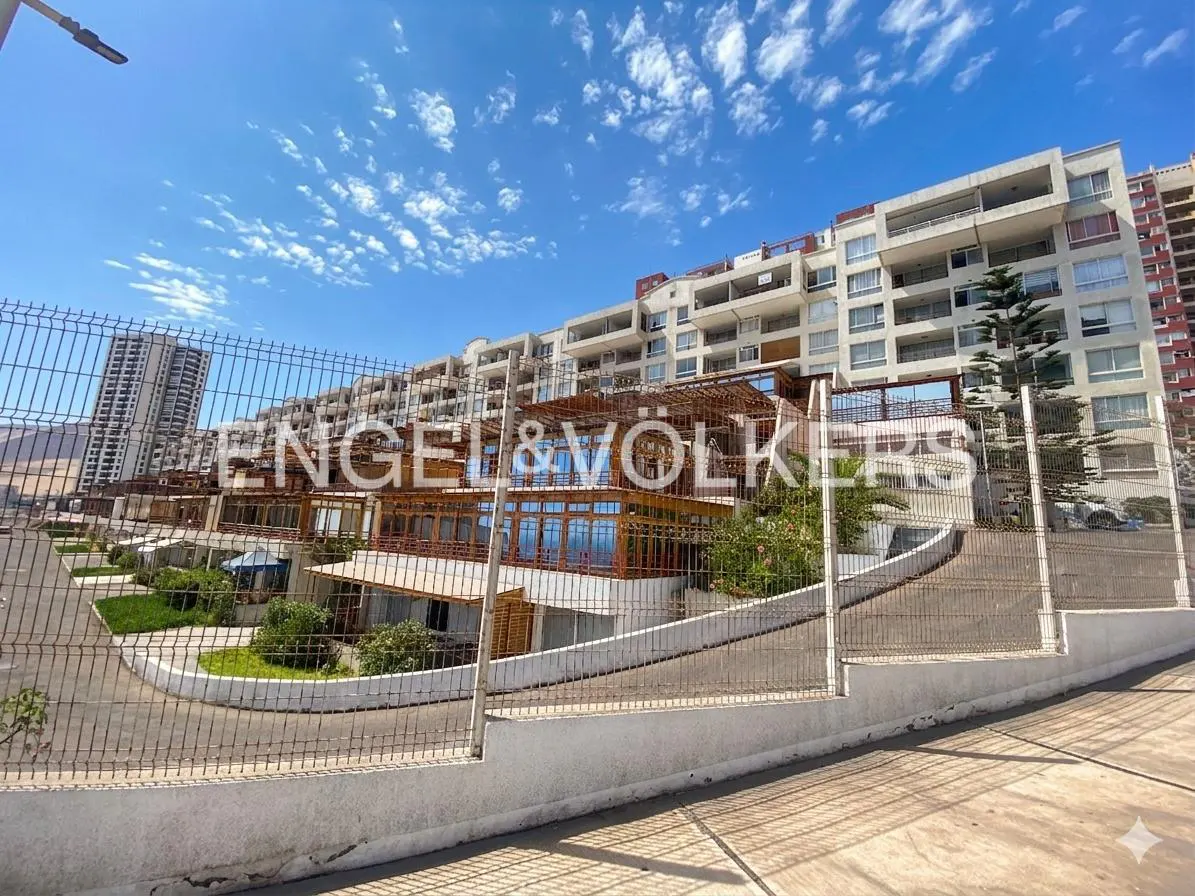 Exterior view of a multi-story, white apartment building with balconies, behind a wire fence, under a blue sky with scattered clouds.