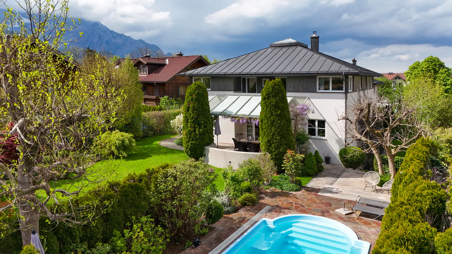 Aerial view of a white two-story house with a gray roof, a blue pool, and a lush green garden.