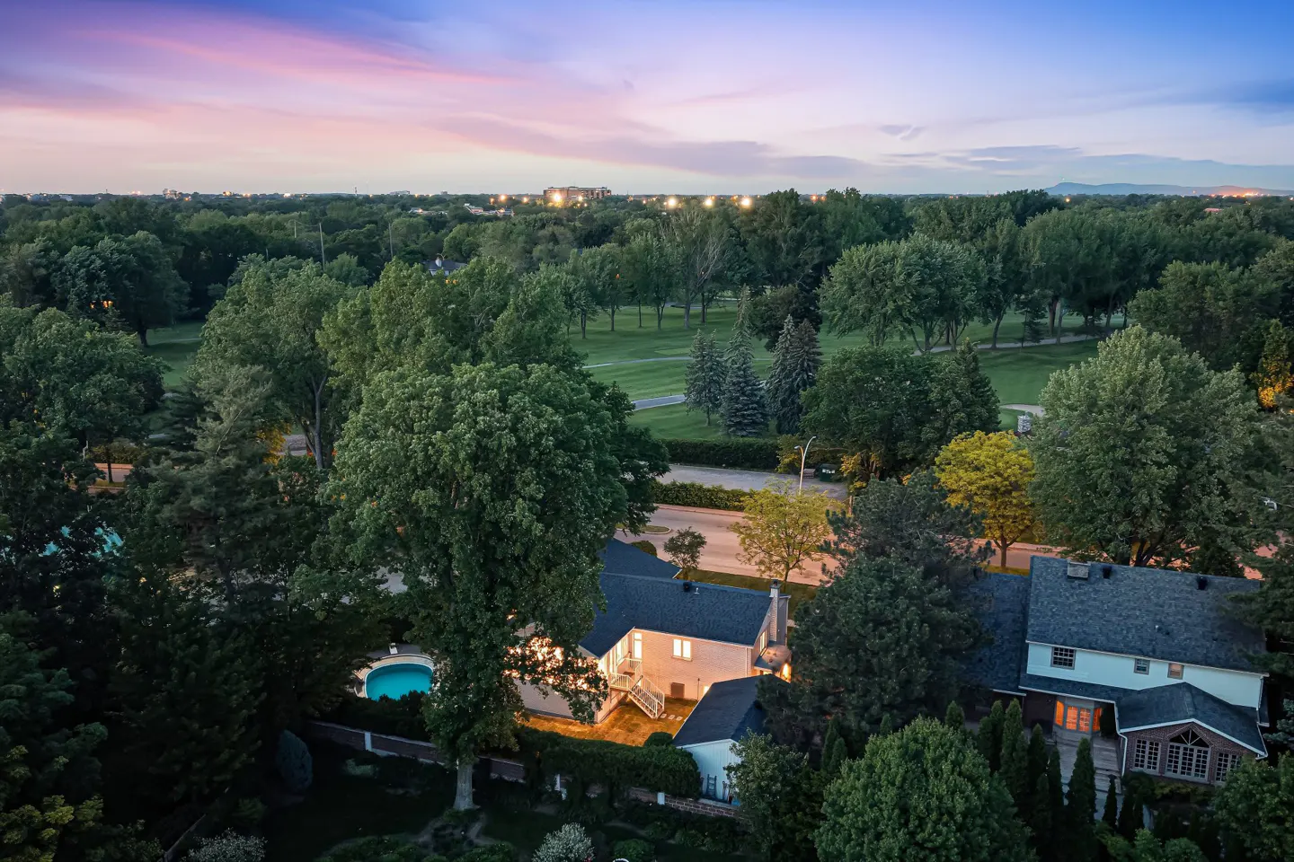 Aerial view of a house with a glowing pool at dusk, surrounded by lush trees and a golf course under a pink sunset sky.