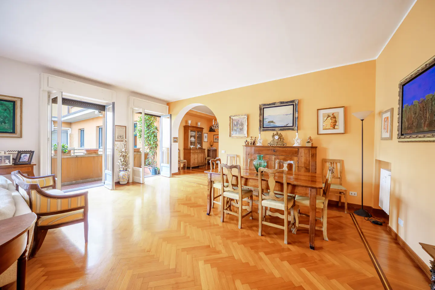Bright, airy living room with wood floors, orange walls, and a balcony. A wooden dining table and chairs sit near a sideboard with artwork above.
