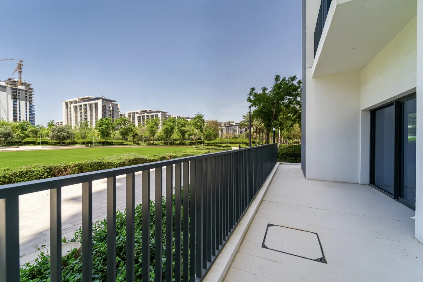 Balcony view of a green lawn, trees, and buildings under a blue sky. The balcony has a gray metal railing and a white floor.