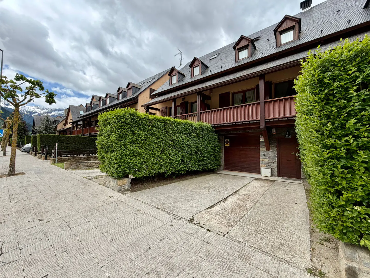 Row of townhouses with brown balconies and dormer windows, separated by green hedges, under a cloudy sky.