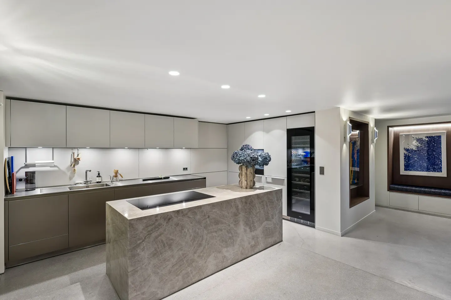 A modern kitchen with gray cabinets, a marble island with a cooktop, and a wine fridge. A blue floral arrangement sits on the island.
