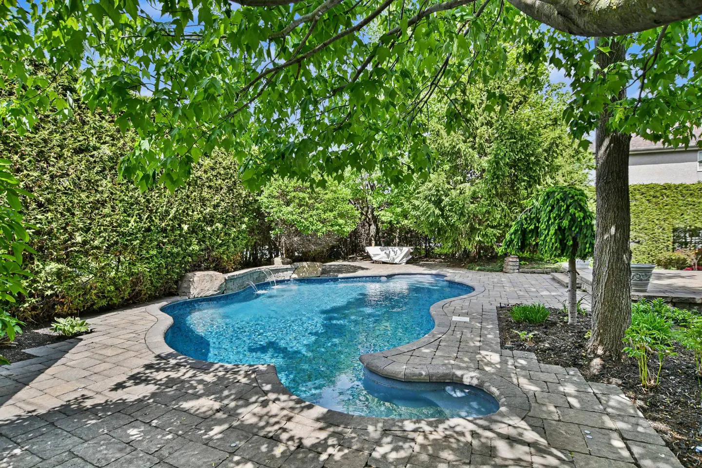 A kidney-shaped pool with blue water is surrounded by a stone patio and lush greenery.