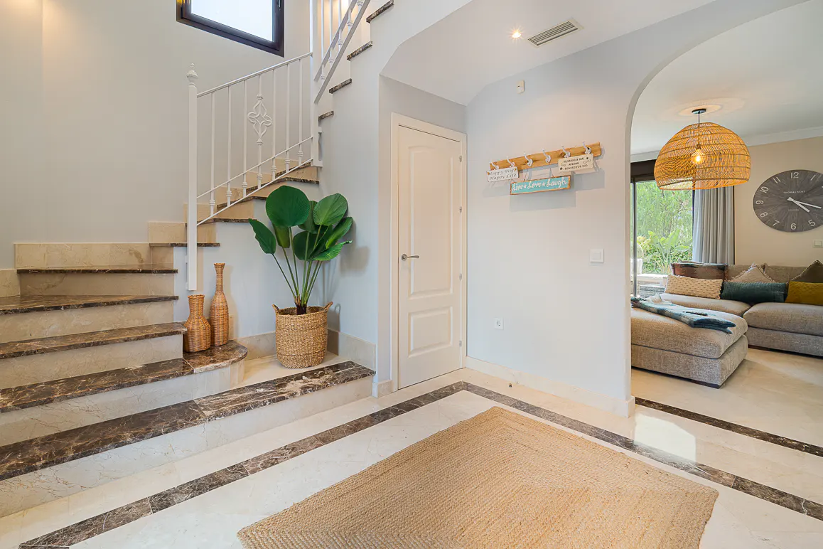 Interior view of a home's foyer with marble stairs, white railings, a potted plant, and a jute rug. A living room is visible through an archway.
