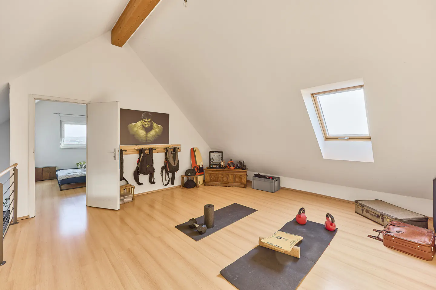 Attic room with wood floors, white walls, and a skylight. Exercise mats, weights, and bags are scattered around. A Hulk poster hangs on the wall.