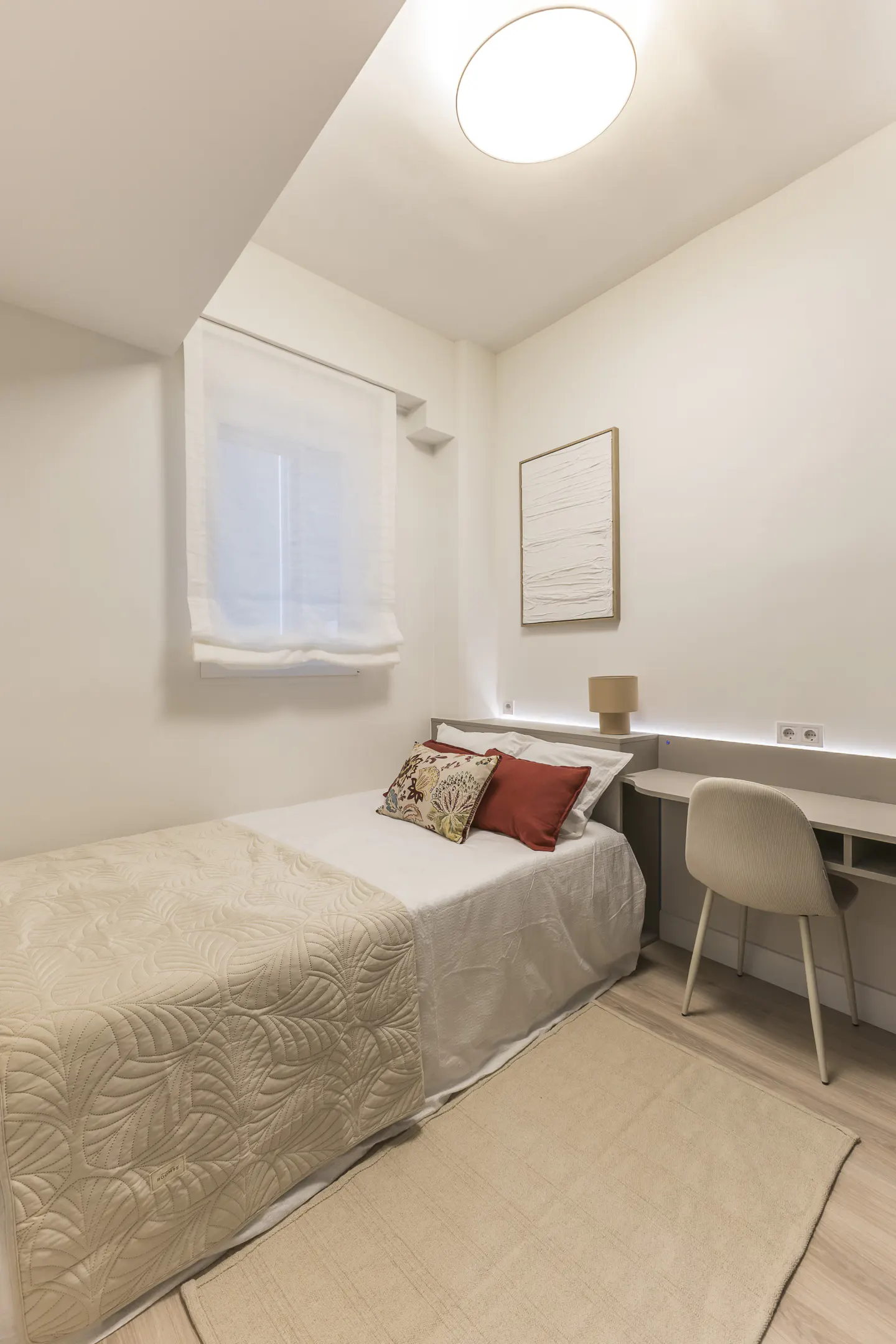 A bright, modern bedroom with a single bed, desk, and chair. The room is painted white with a beige rug and bedding.