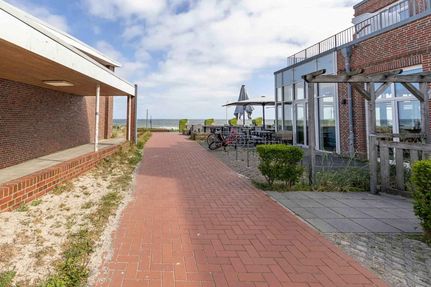 A red brick path leads toward the sea between a modern brick building and a restaurant patio with bicycles and umbrellas.