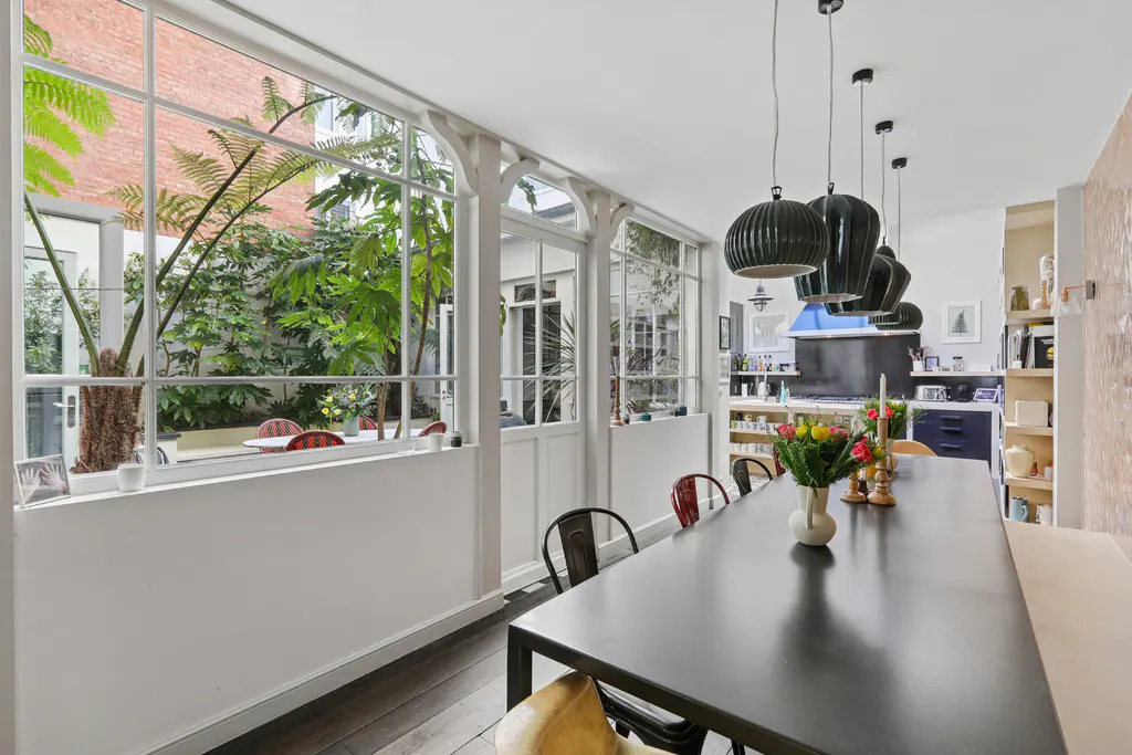 Bright dining room with a long black table, chairs, and pendant lights. A wall of windows overlooks a lush green courtyard.