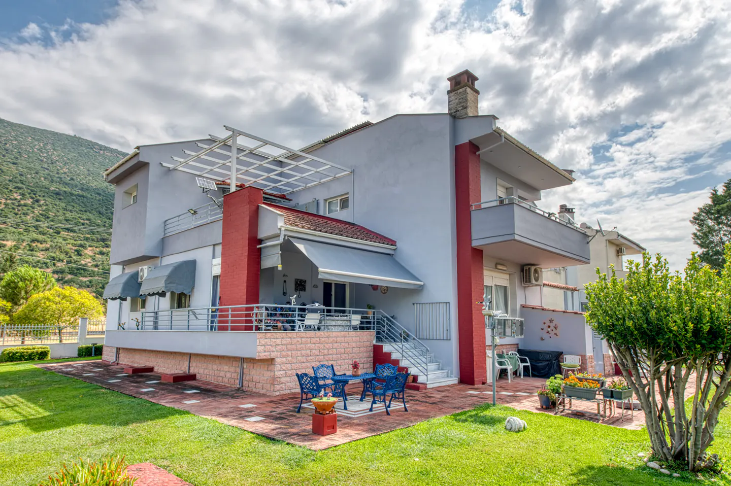 Two-story gray house with red accents, a patio with blue furniture, and a green lawn under a cloudy sky.