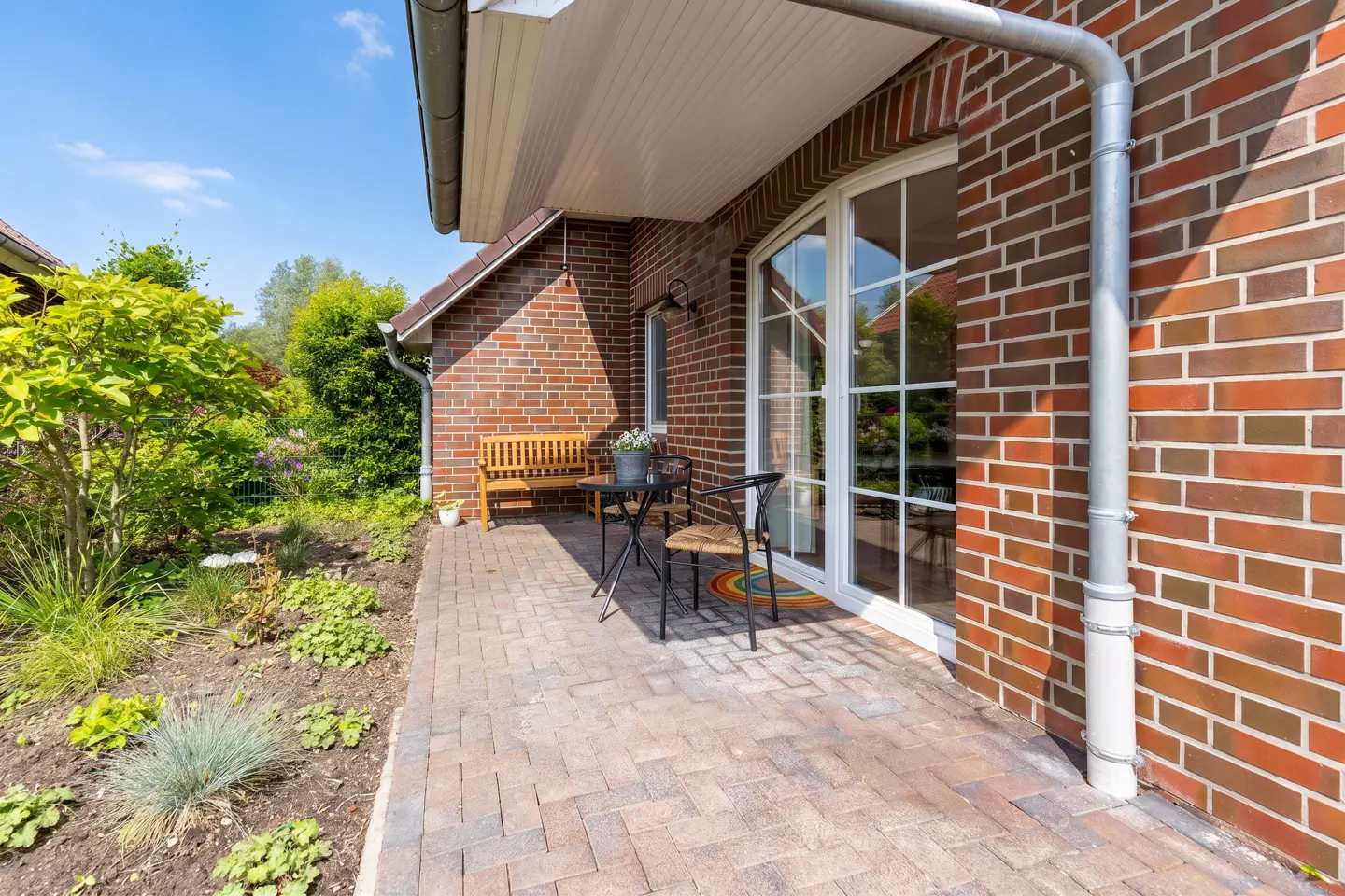 A brick patio with a small table, two chairs, and a wooden bench next to a house with large glass doors and a garden.
