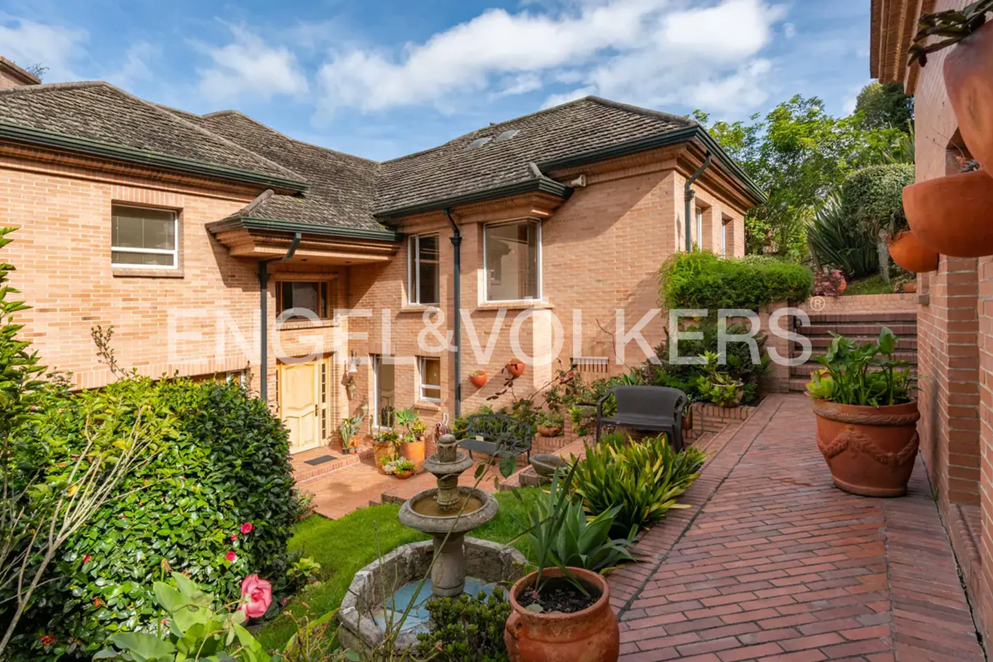 Exterior of a brick house with a fountain, garden, and brick walkway under a partly cloudy sky.
