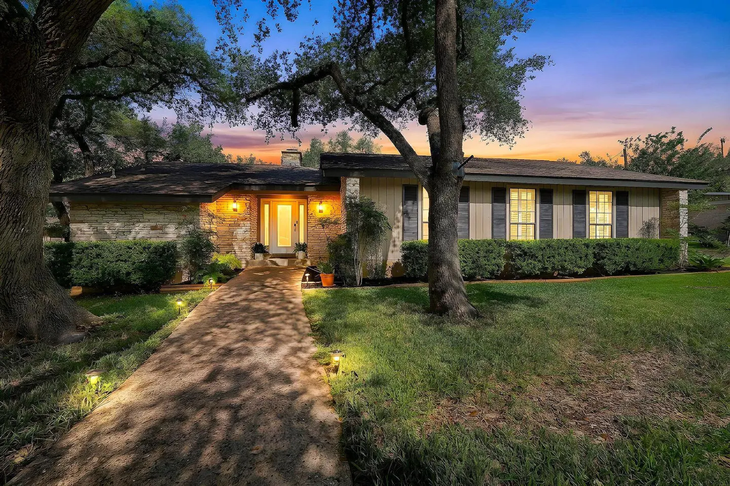 A ranch-style house with a stone and wood facade, illuminated walkway, and green lawn at dusk.